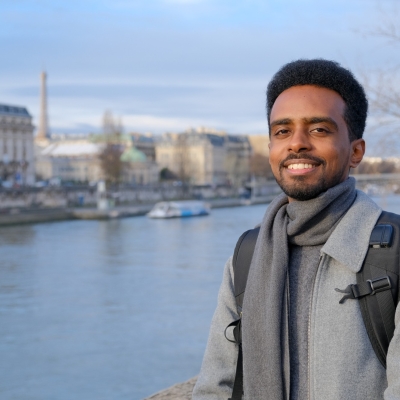 A man standing next to a river. He is smiling at the camera. On the other side of the river are some grand buildings, and in the distance is a boat.