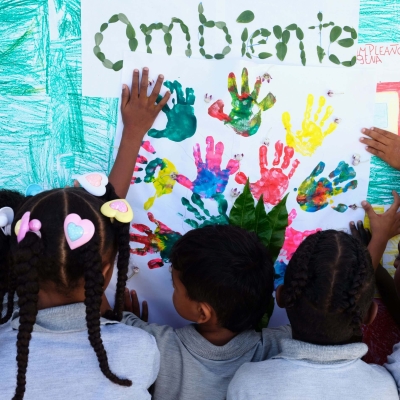 Children painting a picture with their hands. The photo is taken from behind the 5-6 children, and they have made handprints in different colours on a piece of paper, with the word 'ambiente' painted in what looks like fingerprints.