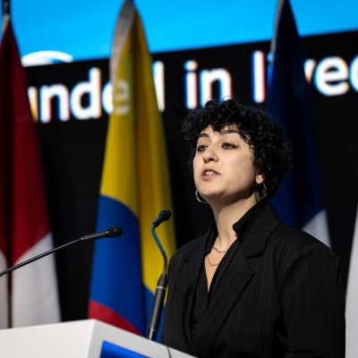 A woman standing at a lectern speaking, there are flags and a large screen behind her