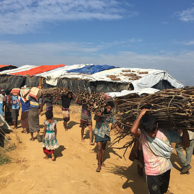 People carrying firewood over their shoulders in a camp setting.
