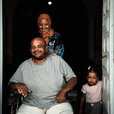 Family in a doorway, man is in a wheelchair with woman standing behind him. They are both laughing. Next to them, a little girl frowns at the camera