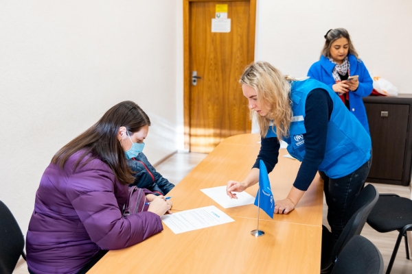 Woman receiving vouchers from UNHCR staff members sits at a table completing forms