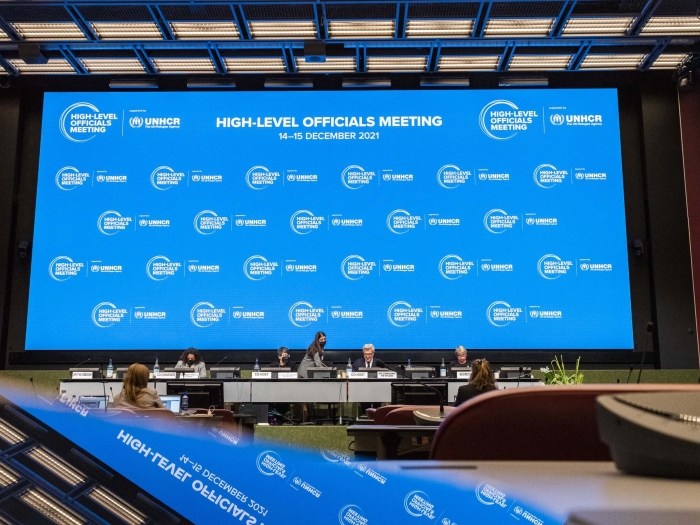 A view from the back of the room towards the stage, where there is a large screen with the High-Level Officials Meeting logo repeated across it. This, and the ceiling, are reflected in the foreground of the image, where a laptop or something is sitting