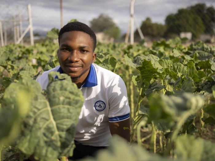 A young man crouching among green plants, smiling at the camera