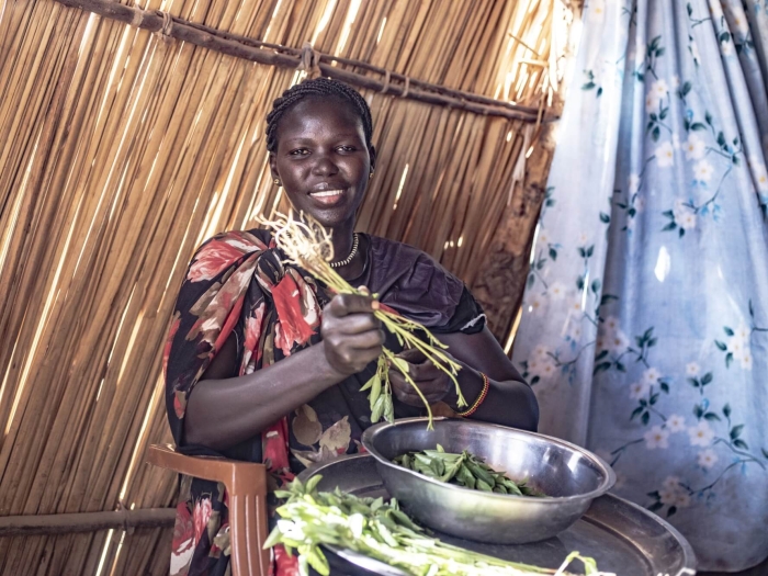 A woman mixing vegetables in a metal bowl which is sitting on a metal tray with more vegetables. The wall behind her is made of straw