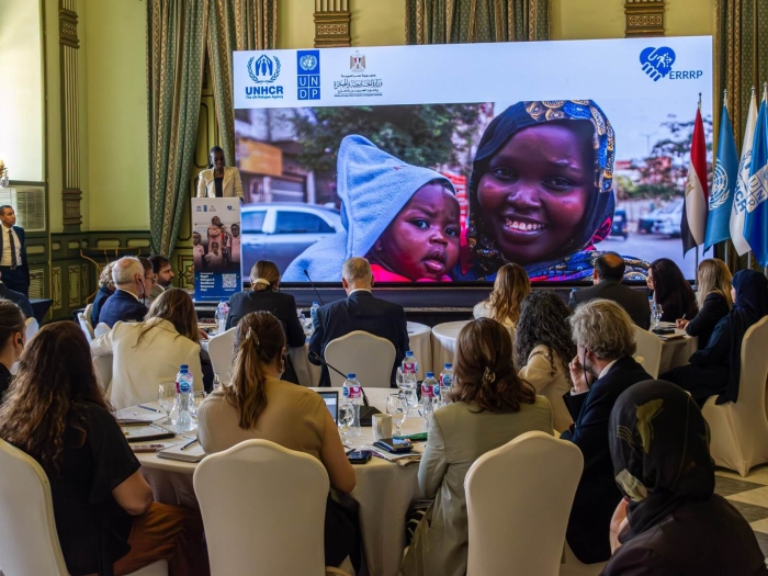An event with people sitting round tables - in the background is a screen with a photo of a woman and child, and someone is standing at a lectern speaking