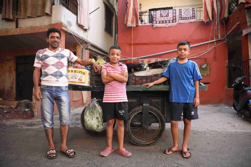 Two boys stand with their father in front of vegetable cart