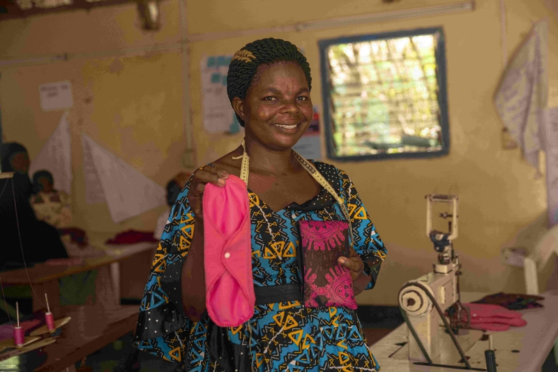 A woman smiling at the camera holds up a reusable pad. There is a sewing machine in the background.