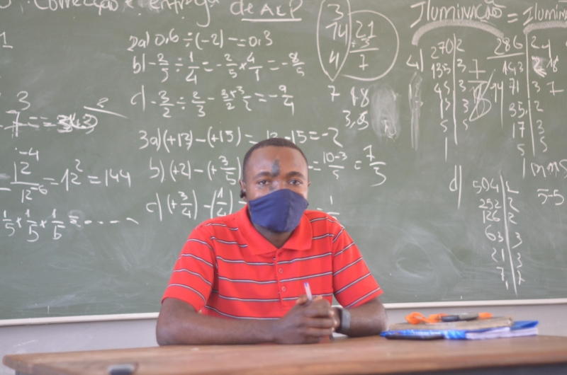 Man sits at a desk with a mask on in front of a black board
