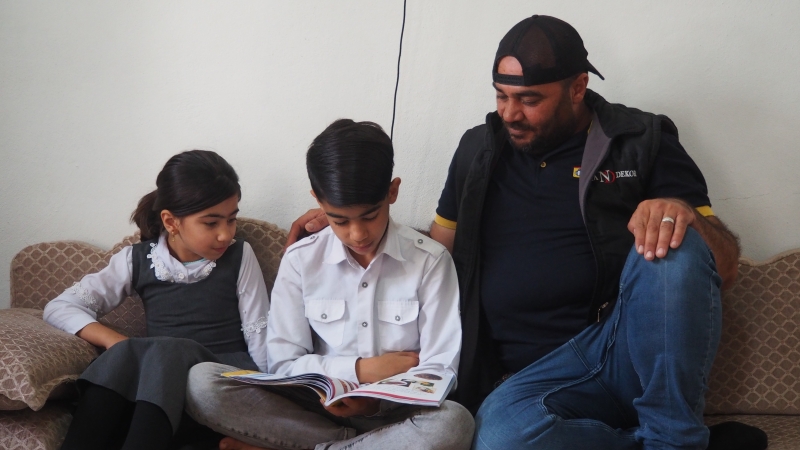 A boy sits in between his sister and father reading a book.