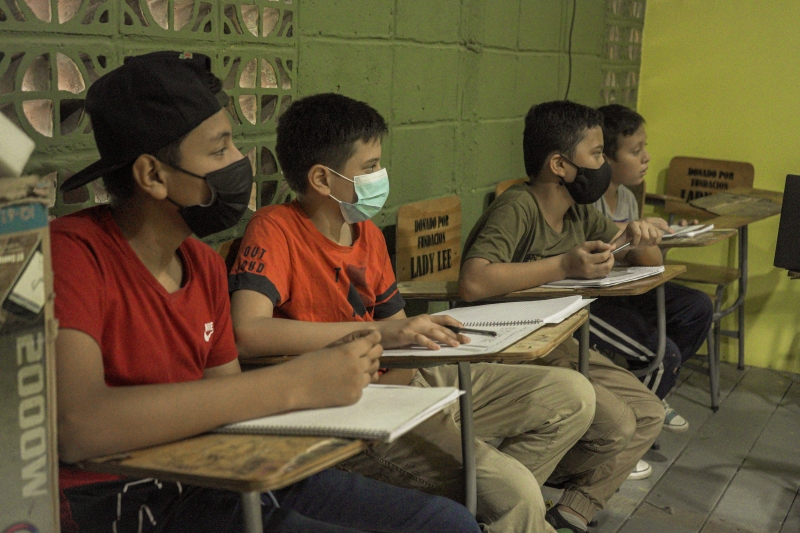 Children wearing masks are sitting a school tables with notebooks on them