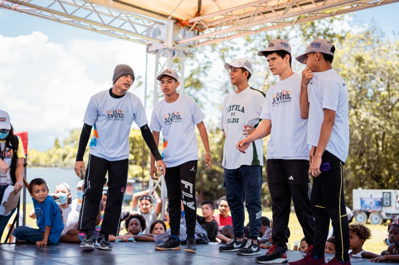 Four boys stand on a stage, a group of children in the background are watching them