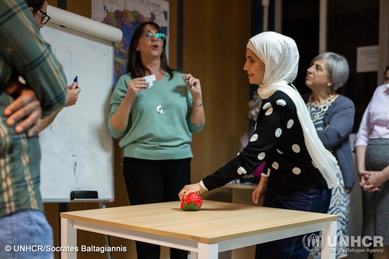 People participating in a naturalization course - a woman has a strawberry-form stress ball in her hand, resting on a table in front of a white board where someone is drawing a grid
