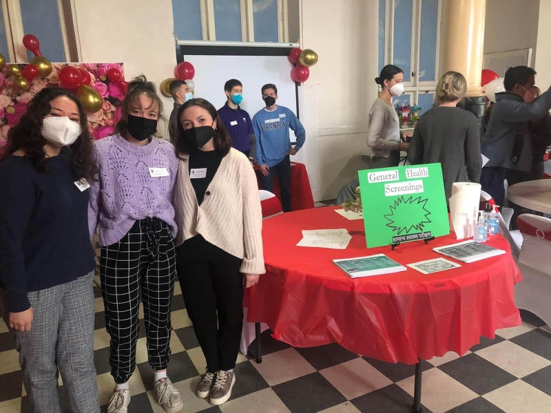 People wearing masks standing facing the camera. They are at a health fair, there is a sign "General Health Screenings" on the table."