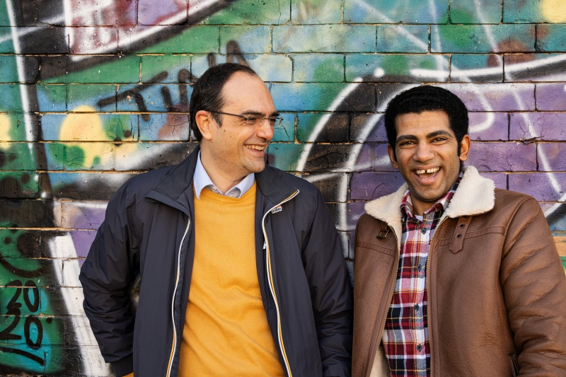 Two men stand in front of a wall with a graffiti