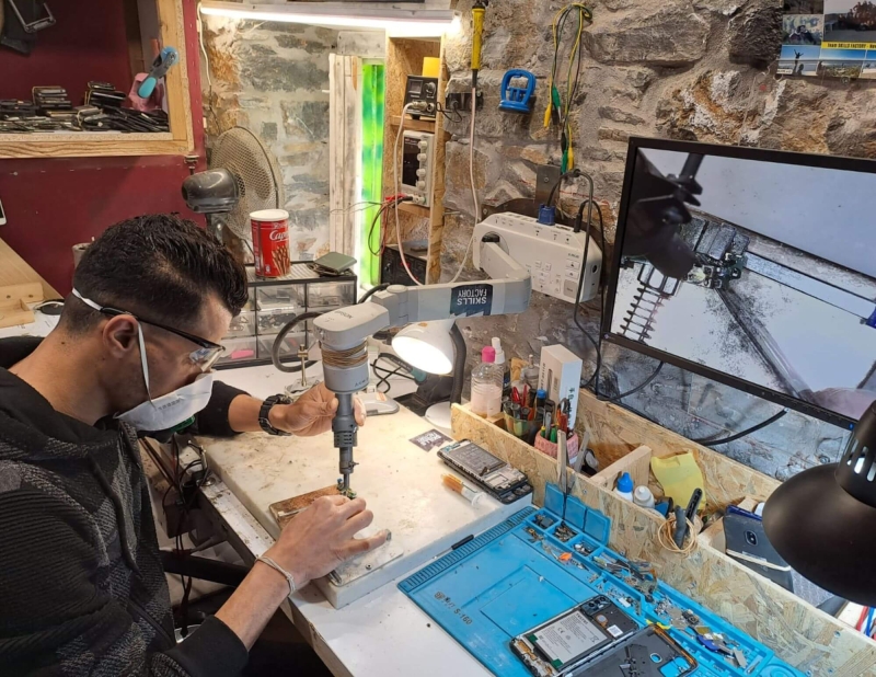 A man in a workshop using a machine - there are tools, tins and a computer around him