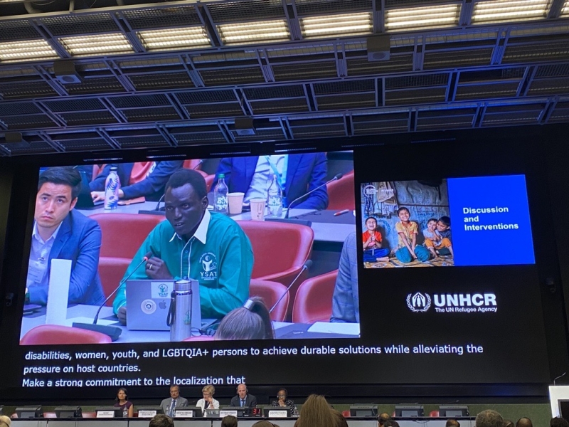 Panel at meeting with a large screen behind them, projecting video of John Jal Dak speaking from the floor.