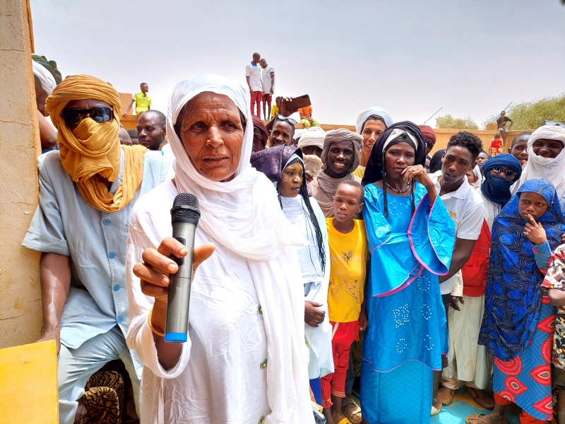 A woman speaks in a microphone, a group in the background listens
