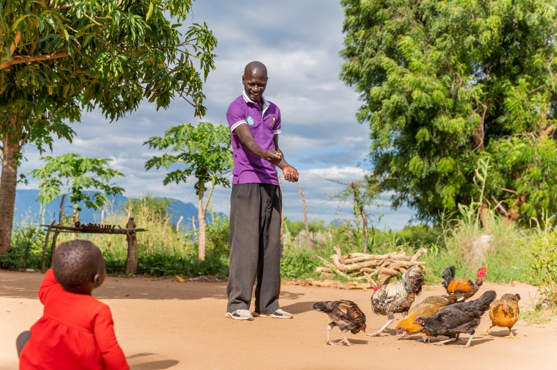 A man smiles at a child sitting on the floor as he tends to his chickens