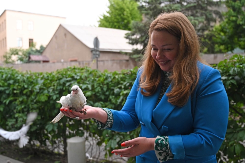 A woman in a blue suit is smiling with a white bird perched in the palm of one hand. She is outside with hedges and buildings behind her and another bird is in flight just behind her.