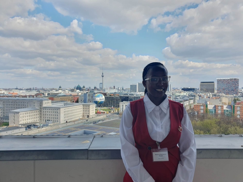 Rebecca standing on the balcony of German Federal Ministry for Economic Cooperation and Development, behind her is the Berlin skyline, with the TV Tower (Fernsehturm) and Berlin Cathedral (Berliner Dom) in the distance and the Welt hot air balloon in the foreground. The sky is blue with white/light grey clouds.