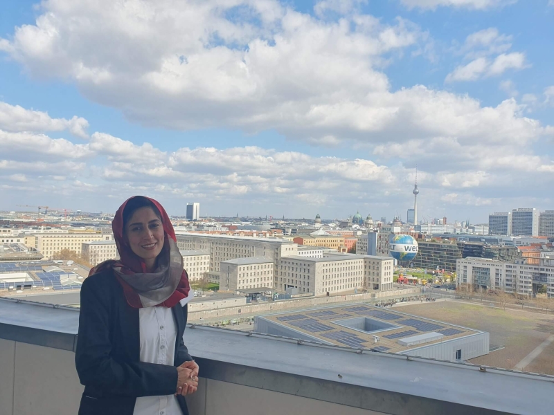 Safia standing on the balcony of German Federal Ministry for Economic Cooperation and Development, behind her is the Berlin skyline, with the TV Tower (Fernsehturm) and Berlin Cathedral (Berliner Dom) in the distance and the Welt hot air balloon in the foreground. The sky is blue with white/light grey clouds.