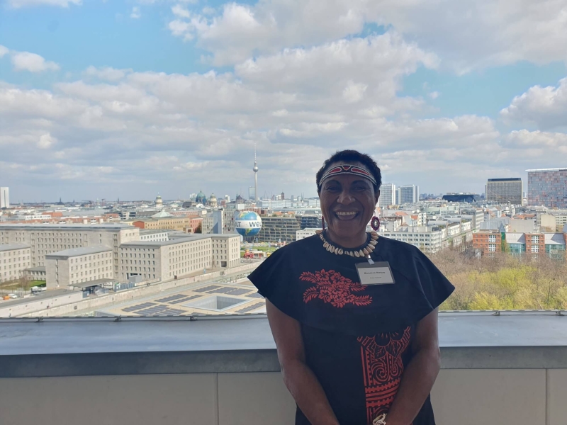 Maryanne standing on the balcony of German Federal Ministry for Economic Cooperation and Development, behind her is the Berlin skyline, with the TV Tower (Fernsehturm) and Berlin Cathedral (Berliner Dom) in the distance and the Welt hot air balloon in the foreground. The sky is blue with white/light grey clouds.