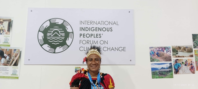 Maryanne sitting in front of a sign saying "International Indigenous Peoples' Forum on Climate Change" with photos on the wall either side of the sign. Maryanne is wearing traditional clothes and a headdress and is smiling at the camera