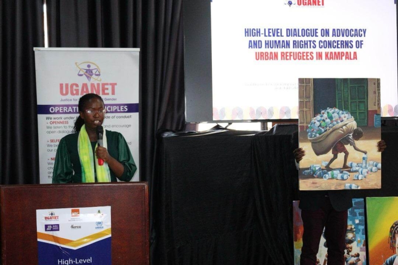 Rebecca standing at a podium and speaking into a microphone at UGANET - High-Level Dialogue on Advocacy and Human Rights Concerns of Urban Refugees in Kampala - next to her someone is holding a large image of a child carrying a huge sack of plastic bottles on their back as they stoop to pick up more