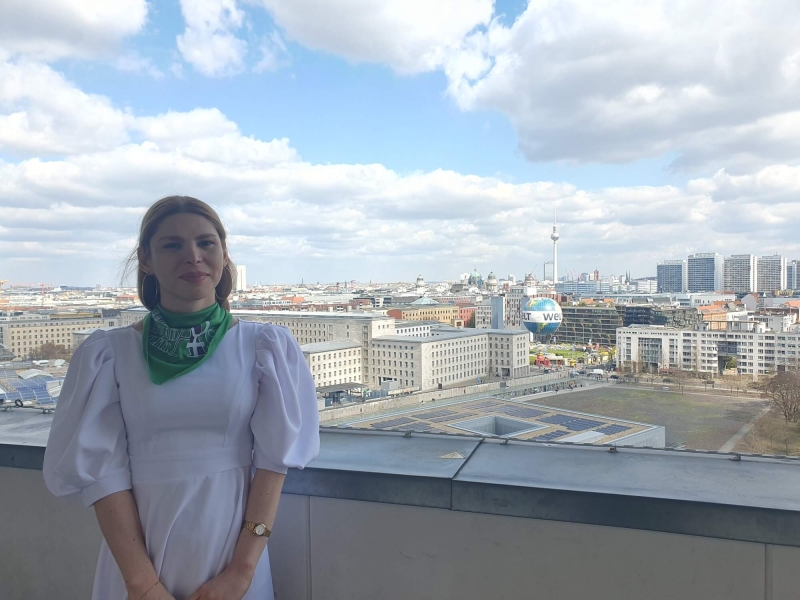 Vlada standing on the balcony of German Federal Ministry for Economic Cooperation and Development, behind her is the Berlin skyline, with the TV Tower (Fernsehturm) and Berlin Cathedral (Berliner Dom) in the distance and the Welt hot air balloon in the foreground. The sky is blue with white/light grey clouds.