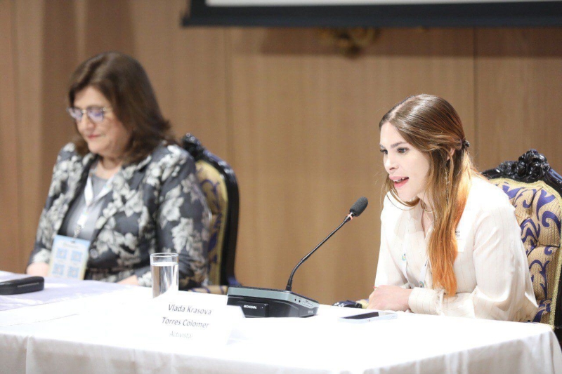 Vlada sitting at a podium with another person next to her. She is leaning forward, speaking into a microphone