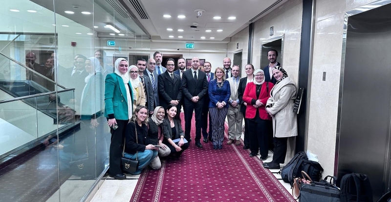 A group photo of 19 people standing in a corridor with a glass wall to the left, lifts on the right, and a red carpet on the floor