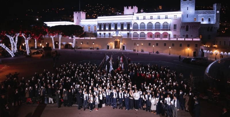 A large crowd of people standing outside a grand building. It is nighttime, the front of the building is lit by lights and the crowd are holding something white in their hands and some flags
