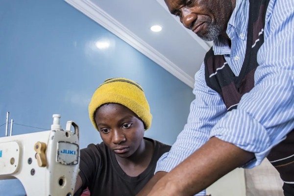 Two people working on a sewing machine, one is sitting behind it, the other is leaning over