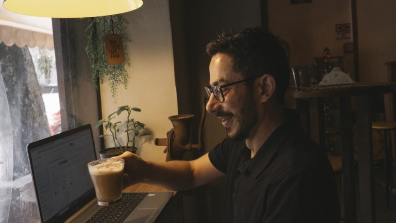 A man raising a cup of coffee in a toast towards his laptop