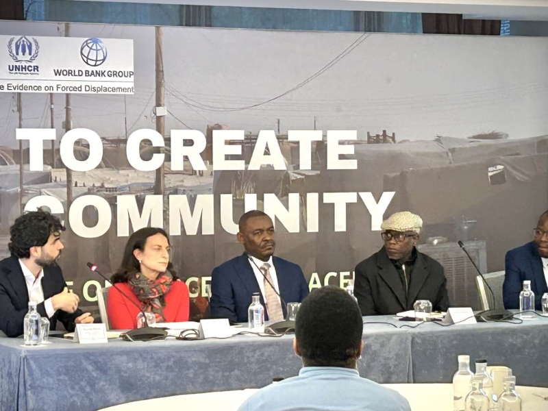 Panel of the event - five people sit behind a table with a backdrop behind them - the text of the backdrop is not really visible.