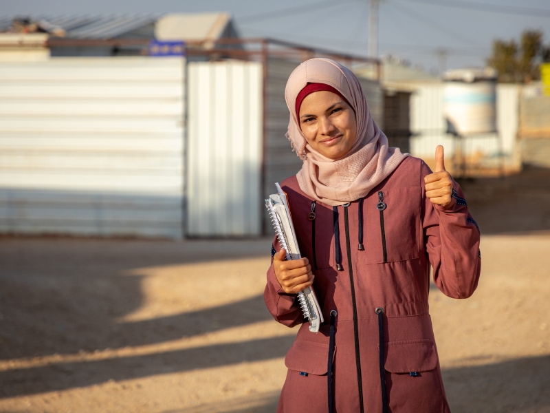 A woman standing outside, with buildings behind her, smiling and giving a thumbs up to the camera. She is holding textbooks and notebooks under one arm