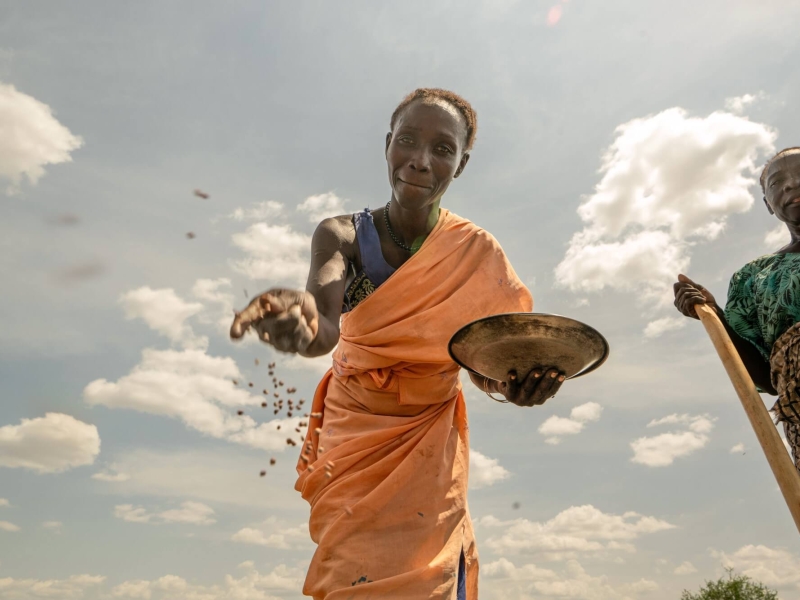 A woman scattering seeds from a metal bowl - the camera is on the ground facing upwards as the seed falls down - the woman is smiling and the sky behind her is bright with a few white fluffy clouds