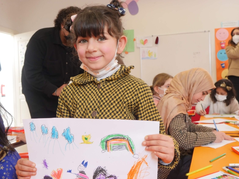 A young girl holding up a colourful drawing of people, rainbows and butterflies and smiling at the camera - there are other people drawing at a table behind her