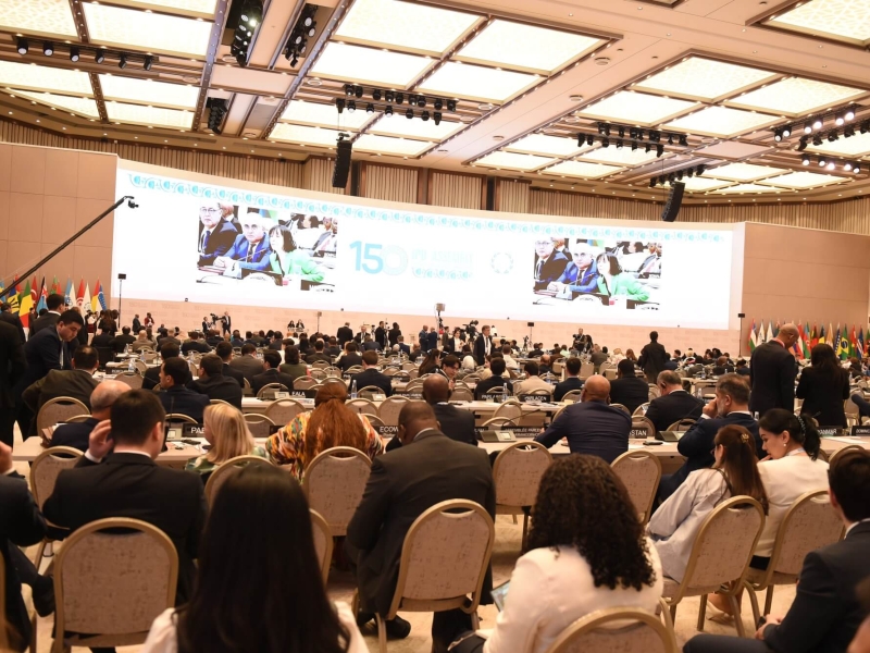 A wide-angle photo of the assembly from behind, showing participants and screens in the distance