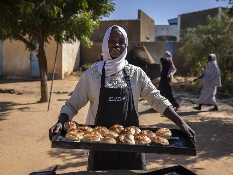 A woman standing holding a tray of baked swirls (cinnamon or raisin type of pastry) - she is wearing an apron, smiling at the camera, standing outside with a tree and some buildings in the background as two people walk behind her