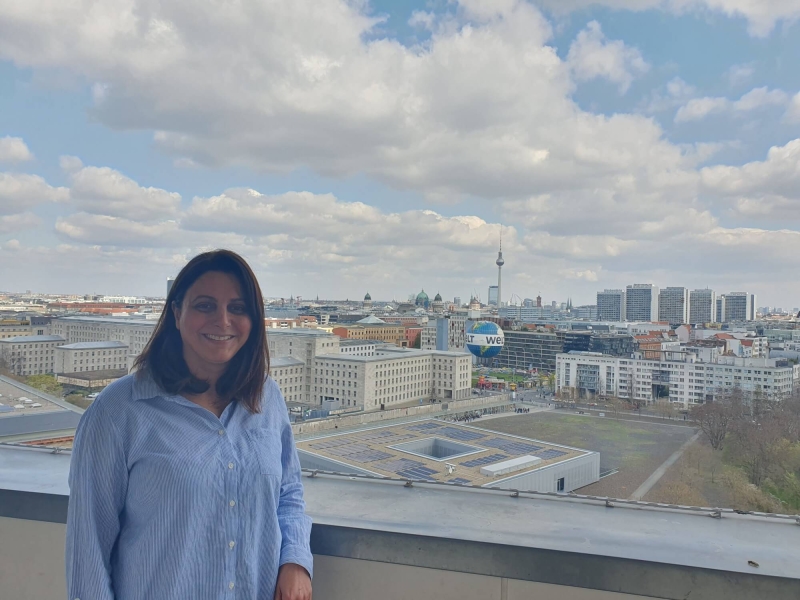 Taban standing on the balcony of German Federal Ministry for Economic Cooperation and Development, behind her is the Berlin skyline, with the TV Tower (Fernsehturm) and Berlin Cathedral (Berliner Dom) in the distance and the Welt hot air balloon in the foreground. The sky is blue with white/light grey clouds.
