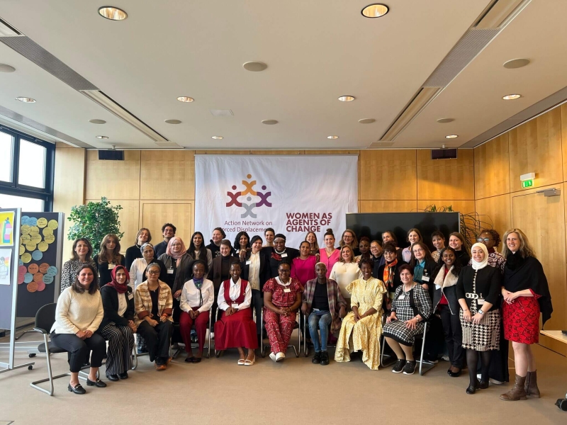 A group photo of participants at the Action Network on Forced Displacement Annual Meeting - around 40 people, those at the front sitting on chairs with others standing behind them. Behind them on a wall is a large banner with the Action Network's logo and the words "Women as Agents of Change"