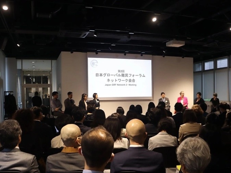 A photo from the meeting, taken at the back of the room, showing participants sitting down, with eight people standing at the front facing them, the lights shining on them, and a screen behind them saying with some writing in Japanese, and in English the words "Japan GRF Network 2nd Meeting"