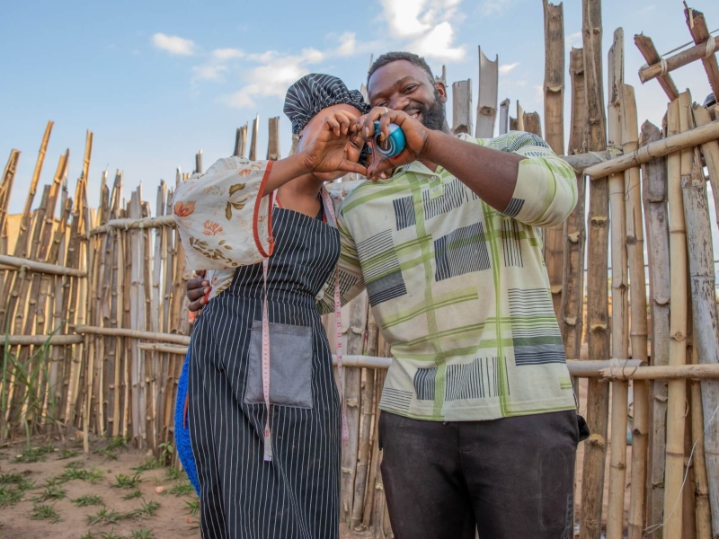 A man and a woman standing together outside, with a wooden fence behind them. They have their arms around one another, and are making a heart shape with his left hand and her right.