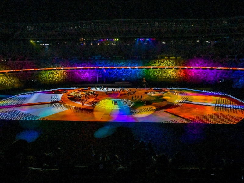 Inside the stadium, it is dark, except for the centre which is lit up with a lot of different coloured bright lights. In the middle, there are people on the stage, and the stands are filled with crowds