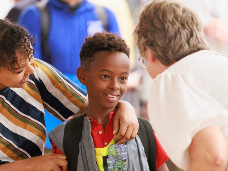 A boy being greeted by a woman, who is bending down to speak to him. He is holding a bottle of water upside down, smiling, while his mother has her arm around his shoulders and is looking at him