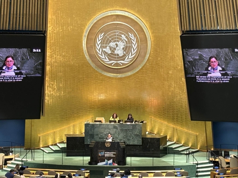 The Mexican Minister delivering the statement at the UN headquarters in New York. She is standing on a stage, with a large golden wall behind her with the UN logo on it. To the sides of the wall are large screens showing her speaking.