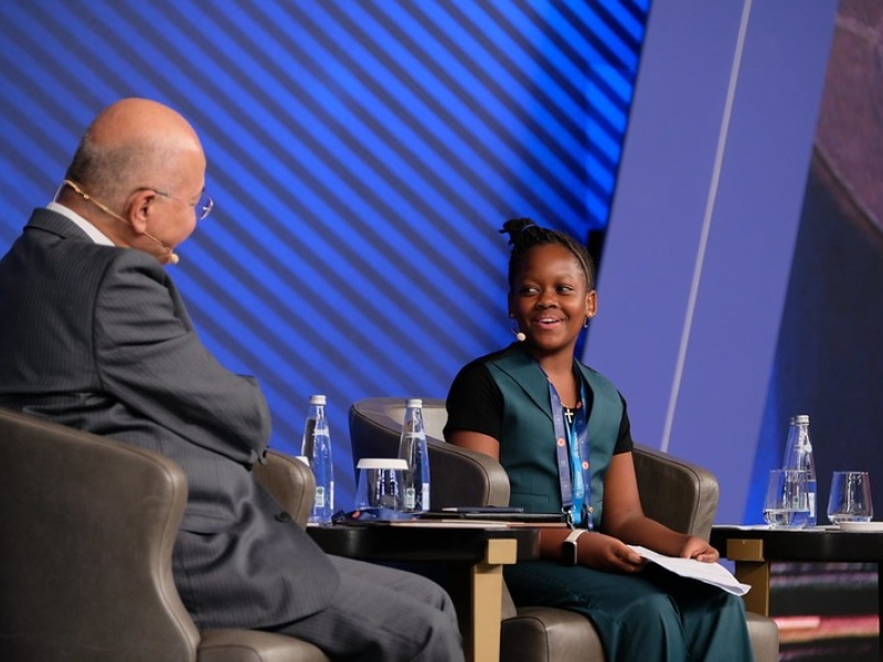 The High Commissioner and Ms. Magesa sitting in chairs on stage. The photo is taken from the side, showing the side and back of the High Commissioner, and the face of Ms. Magesa. She is smiling.