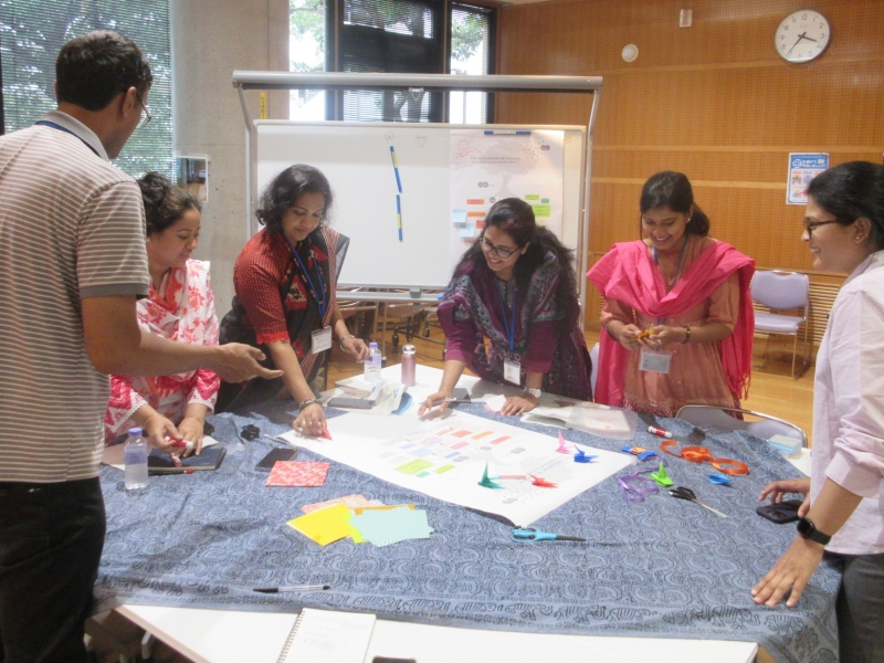 A group of six people standing round a table putting colourful pieces of paper (perhaps some origami?) on a large sheet of paper
